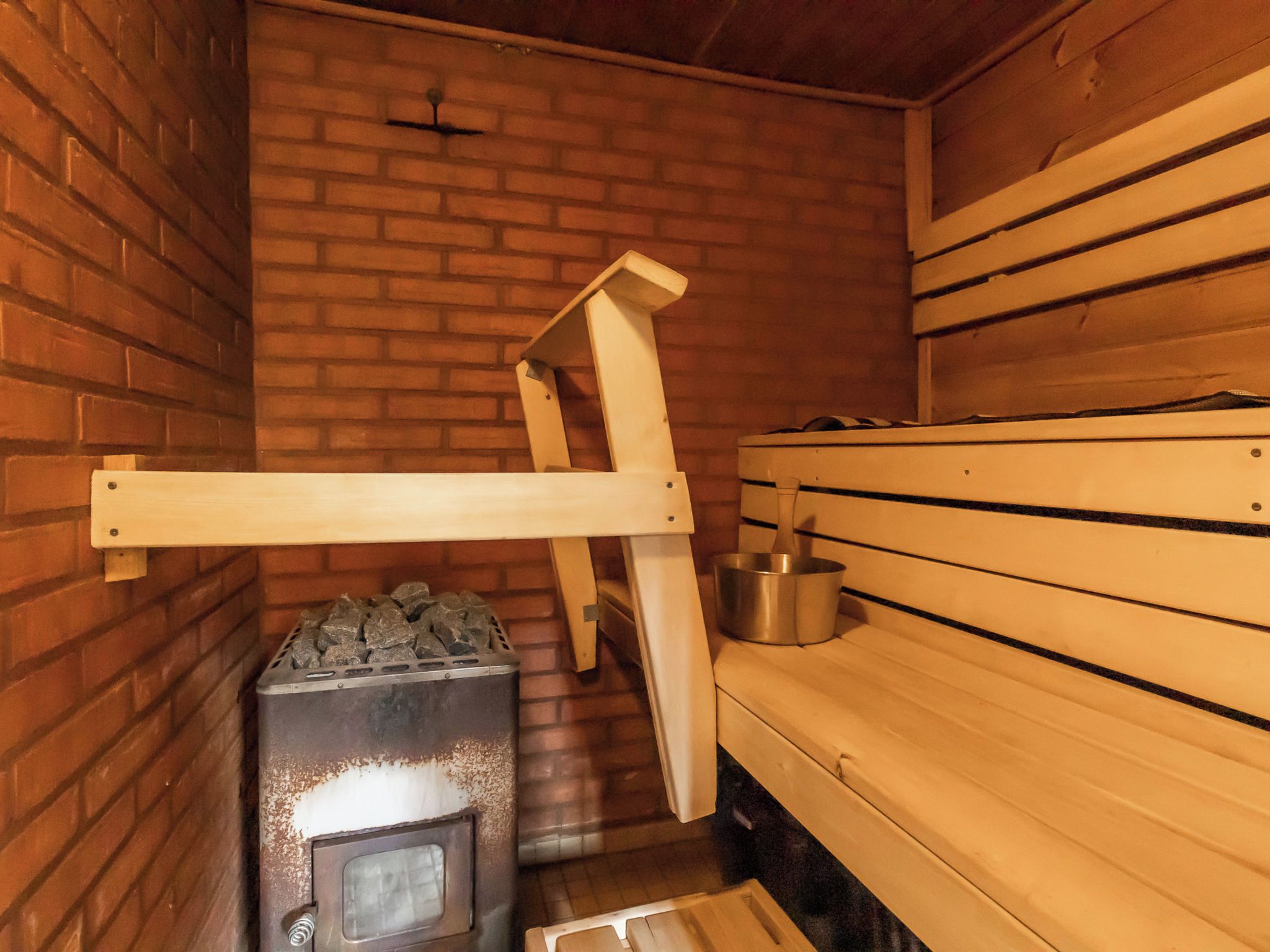 A sauna room with a brick wall, a wooden bench, a sauna heater, and a metal bucket.