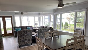 A spacious living area with a dining table, patterned chairs, a ceiling fan, and large windows offering a view of the outdoors. - Douglas Lake House: Summer Days Made for Swimming, Kayaking, and Starry Nights (Levering)