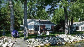 A house with a wooden deck leading to a pond, surrounded by trees and a stone wall. - Douglas Lake House: Summer Days Made for Swimming, Kayaking, and Starry Nights (Levering)