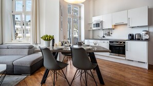 Een moderne keuken met witte kasten, een magnetron en een koffiezetapparaat. Er is een eettafel met stoelen en een grijze bank.