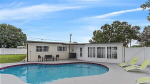 A swimming pool with a curved edge, a covered patio area with a table and chairs, and a fenced backyard. - Florida Living (Bradenton)