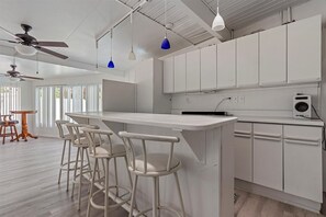 A modern kitchen with white cabinets, a central island with bar stools, pendant lights, and ceiling fans. - Florida Living (Bradenton)