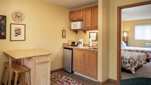 A compact kitchen with wooden cabinets, a microwave, and a coffee maker. There is a small dining table and two stools. The room has a bed with floral bedding and a window with blinds. - Cozy condo w/great location by Flathead Lake - Cozy Marina Cay Getaway (Bigfork)