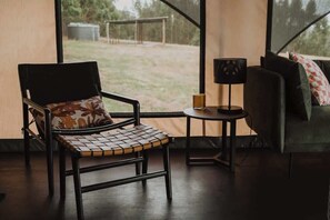 A room with a chair, a small table with a lamp, and a couch with a view of the outdoors.