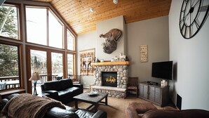 A cozy living room with a stone fireplace, leather sofas, and a large window overlooking a deck.