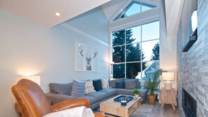 Ein modernes Wohnzimmer mit Ledersessel, grauem Sofa und Couchtisch. Ein Kamin, ein großes Fenster mit Blick auf Bäume und ein wandmontierter Fernseher sind ebenfalls vorhanden.