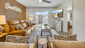 A modern living room with a leather sofa, a coffee table, and a TV. There is a ceiling fan, a neon sign, and a view of buildings through large windows. - NEW Updated 2/2, Beachfront with a private balcony, Atlantic Ocean views, and a heated pool (New Smyrna Beach)