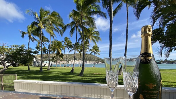 A bottle of Meadowcroft Blanc de Blancs and two glasses on a table with a view of a beach and palm trees. - Waterfront Spacious Home near Beach & Hiking. Family Friendly w/Boat Dock & Pool (Honolulu)
