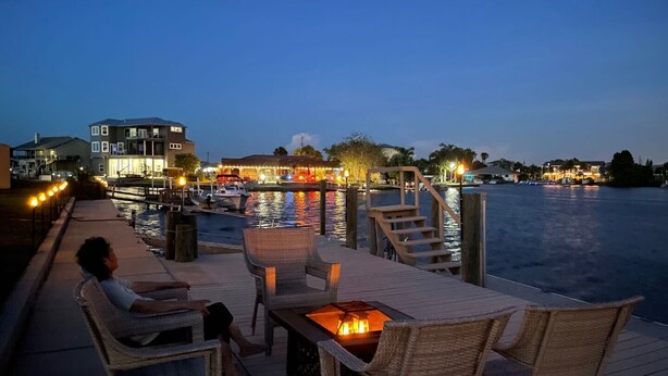 A waterfront deck with wicker furniture and a fire feature, overlooking a marina with boats and buildings. - Seascape - WHOLE HOUSE 360 water views, pool, gulf (Hernando Beach)