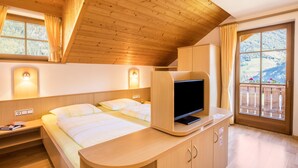 Una habitación con una cama, un televisor sobre un mueble de madera, una ventana con cortinas y vistas a un paisaje montañoso.