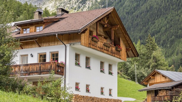 Un chalet en bois à deux étages, doté de balcons et d’une fondation en pierre, avec une chaîne de montagnes en arrière-plan.