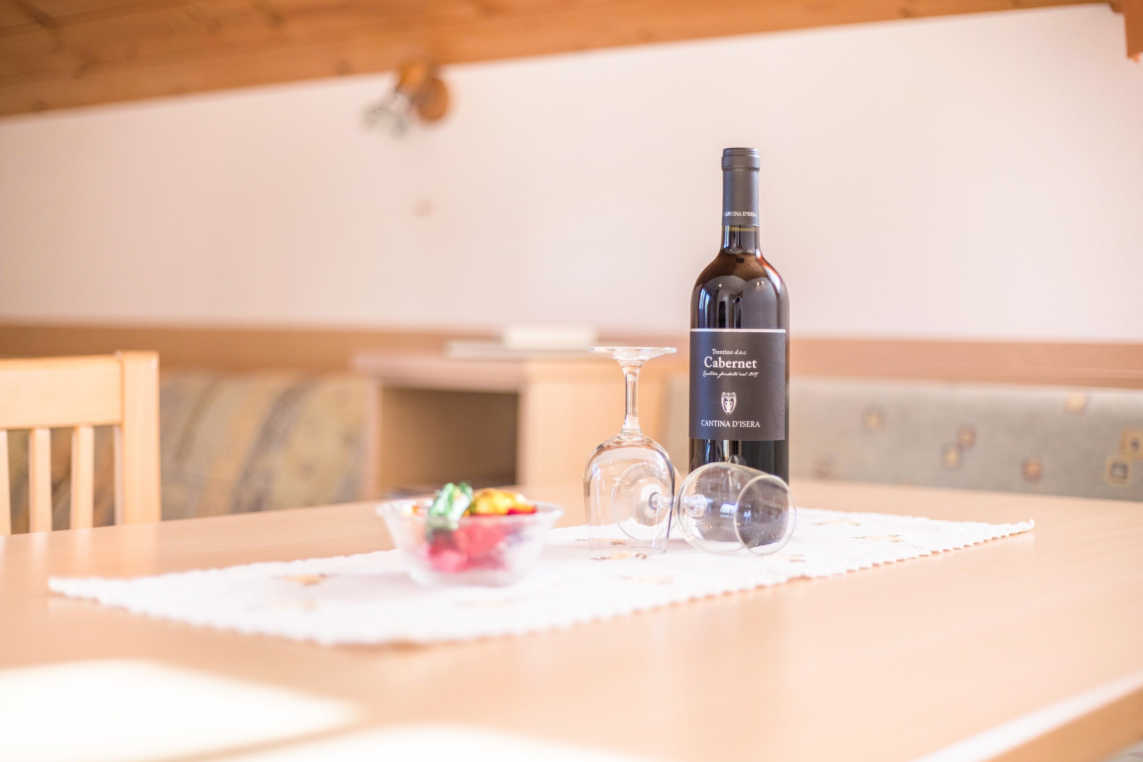A wooden table with a bottle of wine, two wine glasses, and a small bowl of fruit.
