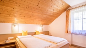 Une pièce avec un plafond en bois incliné, deux lits avec du linge de lit blanc, une table de chevet avec un livre et une lampe, et une fenêtre avec des rideaux.