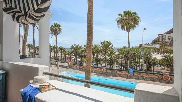 A balcony with a view of a pool, palm trees, and a distant building.