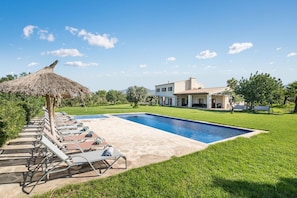 Une piscine avec des chaises longues, un parasol en chaume et une grande maison en arrière-plan.
