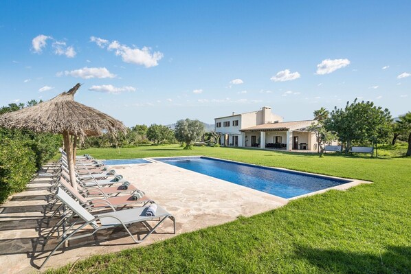 Una piscina con sillas de descanso, una sombrilla de paja y una casa grande al fondo.