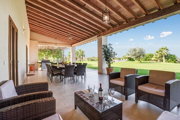 Un espace extérieur abrité, aménagé pour les repas, avec des meubles en osier, une table avec des verres à vin et une vue sur un paysage verdoyant.