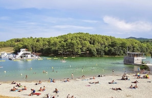 A beach with people sunbathing and swimming, a boat docked on the water, and a forested hill in the background.