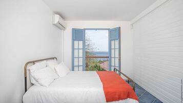 Une chambre avec un lit, des murs blancs, une vue sur l’extérieur à travers des volets ouverts et un balcon.