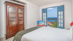 A bedroom with a bed, a wooden wardrobe, a television, and a balcony with blue shutters. - Villa Sol y Sombra the Cuban spirit (Schoelcher)