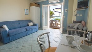 A room with a blue sofa, a dining table with chairs, a TV mounted on the wall, and a view of the outdoors through a door. - Punta Contessa - Punta Contessa n°4 (Capoliveri)