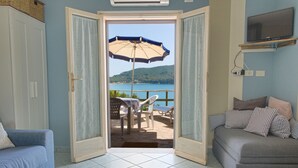 A room with a view of a lake, a sofa, a chair, and an open door leading to an outdoor seating area with a table and umbrella. - Punta Contessa - Punta Contessa n°4 (Capoliveri)