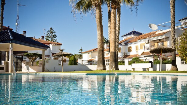 A swimming pool with clear blue water, surrounded by palm trees and white buildings with red-tiled roofs. - Apartment Casa Titan (Riomar) (Pilar de la Horadada)
