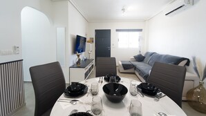 A dining area with a round table set for four, a grey sofa, and a TV mounted on the wall. - Apartment Casa Titan (Riomar) (Pilar de la Horadada)