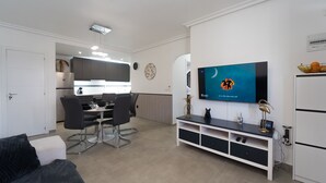 A modern living room with a flat-screen TV, a dining area with a table and chairs, and a kitchenette in the background. - Apartment Casa Titan (Riomar) (Pilar de la Horadada)