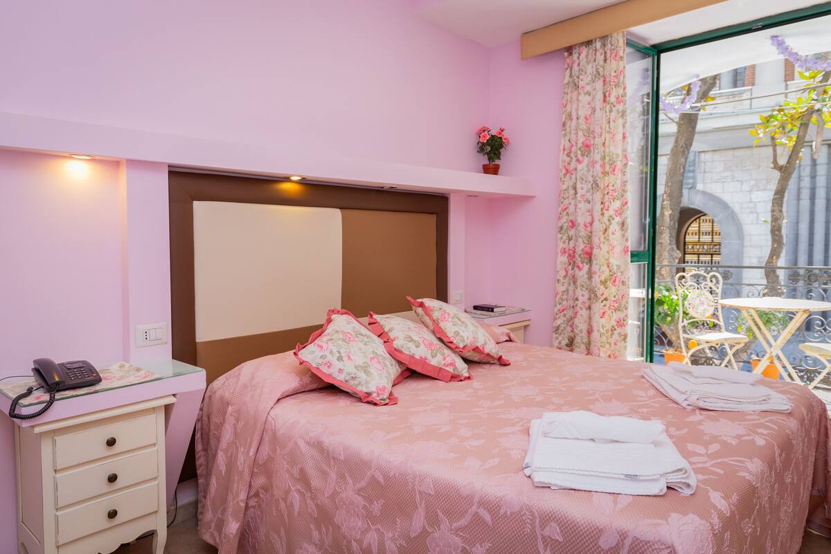 A bedroom with a bed, bedside table, and a small table with chairs outside the window.