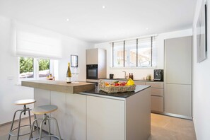 A modern kitchen with a central island, bar stools, a wine bottle, and a fruit basket.