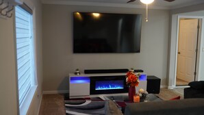 Un salon doté d’un téléviseur à écran plat fixé au mur, d’une cheminée avec un éclairage bleu, d’un ventilateur de plafond et d’une porte donnant sur une autre pièce.