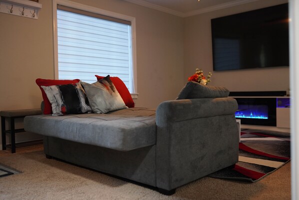 A grey sofa with red and black cushions, a flat-screen TV mounted on the wall, and a fireplace with a blue flame. - "The Little Grey House" (Columbus)