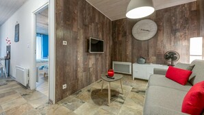 A living room with a wooden wall, a sofa with red cushions, a small table with a red vase, a clock, and a television.