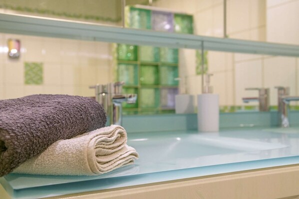 A bathroom with a glass countertop, a folded towel, and a mirror reflecting a green-tiled wall and a sink.