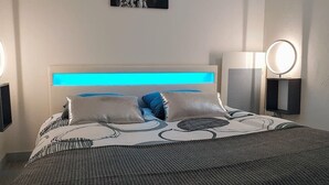 Una cama bien hecha con cabecera que tiene una tira de luz azul, dos almohadas y una manta gris. Hay una mesita de noche con una lámpara y un espejo redondo.