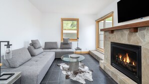 Una sala de estar moderna con chimenea de piedra, un sofá gris, una mesa de centro de vidrio y un televisor de pantalla plana montado en la pared.