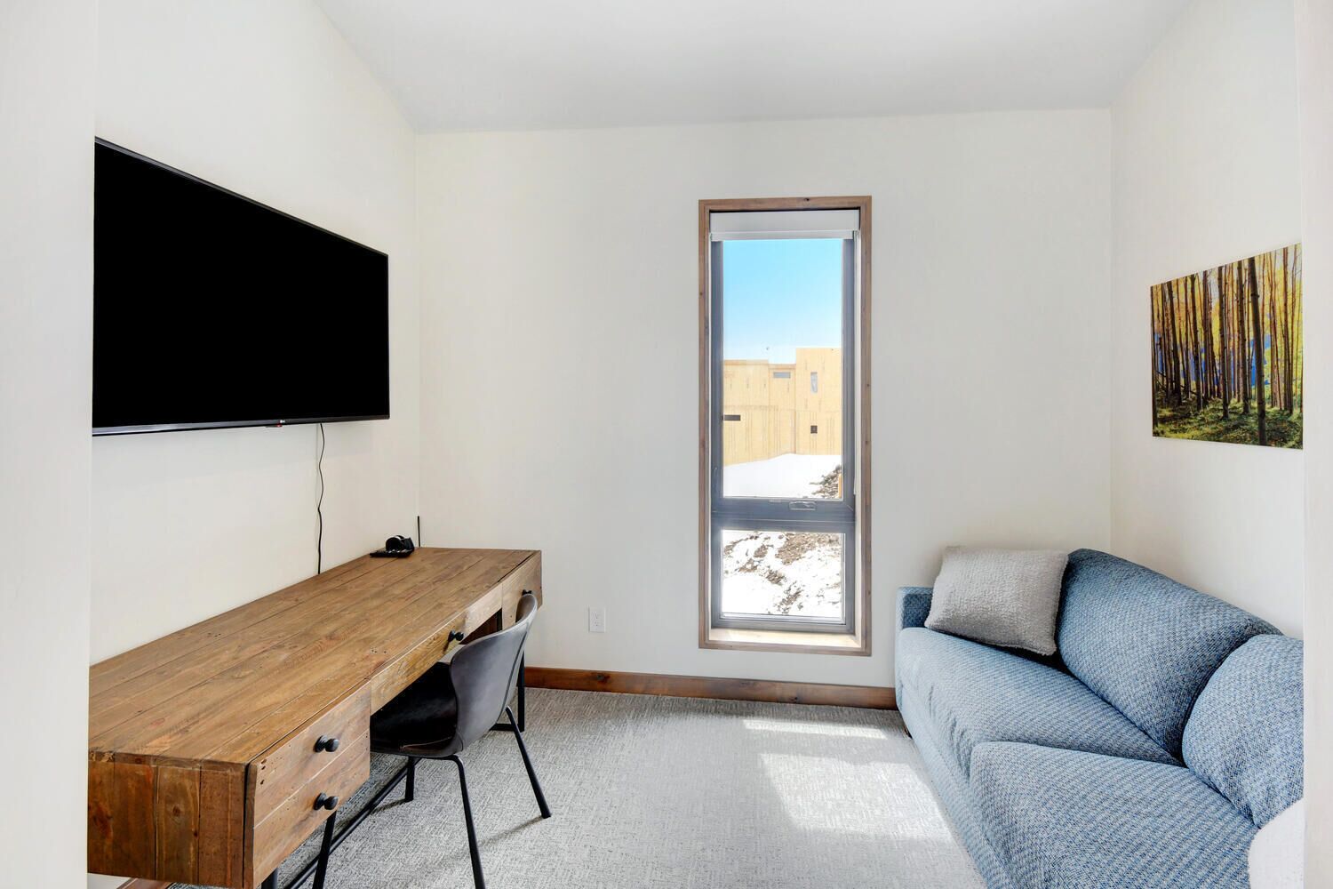 A modern room with a wooden desk, a grey chair, a blue sofa, a flat-screen TV mounted on the wall, and a large window with a view of a building and snow outside.