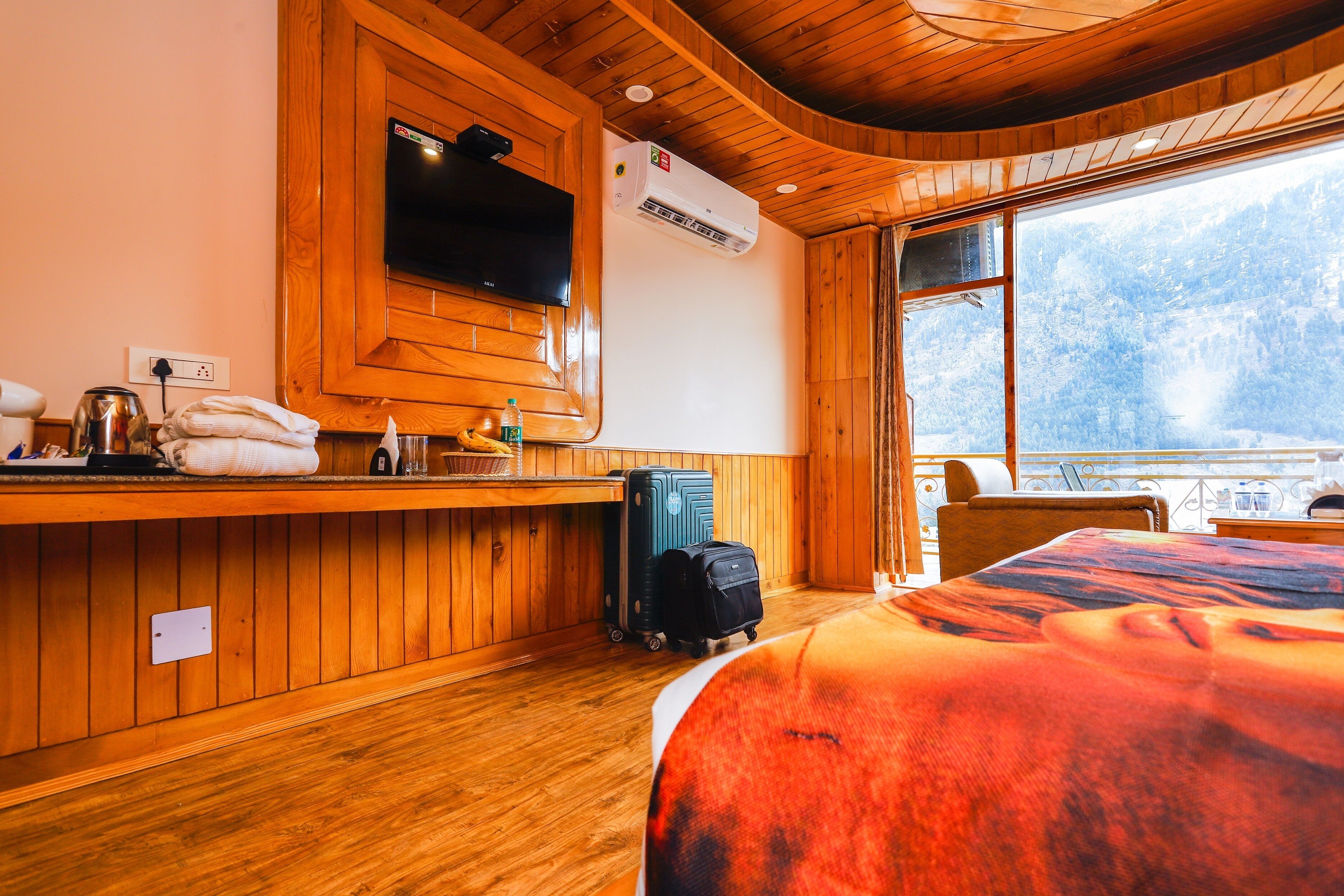 A wooden cabin room with a bed, a TV mounted on the wall, a window with a view of trees, and a suitcase on the floor.