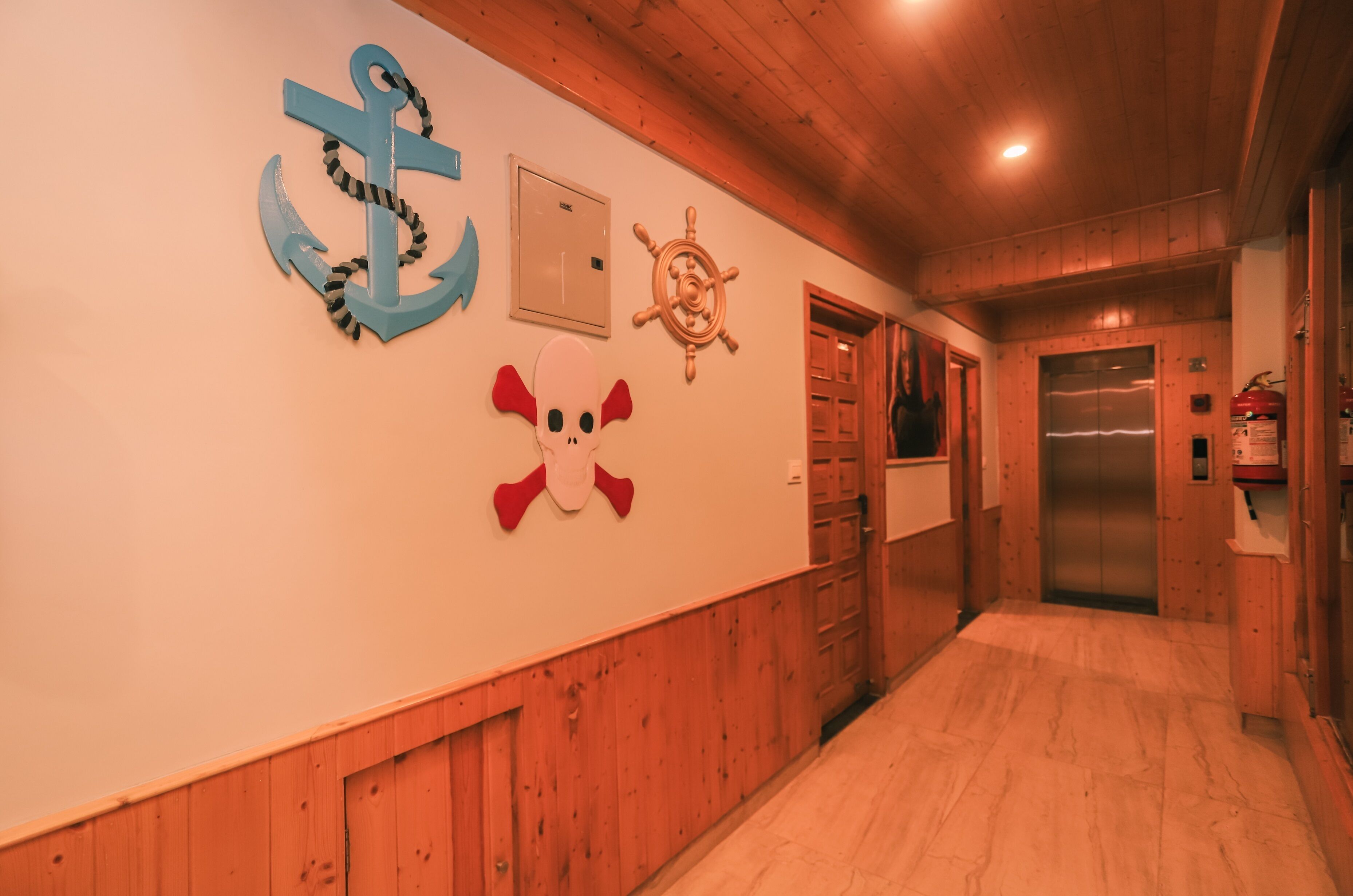 Ein Flur mit Holzwänden und Holzboden. Maritime Dekorationen schmücken die Wand, darunter ein Anker, ein Steuerrad und ein Schädel. Im Hintergrund ist ein Aufzug zu sehen.