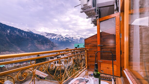 Un balcon donnant sur des montagnes enneigées, une table avec un livre et une bouteille de vin, et une porte en bois.