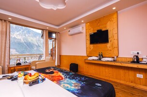 Habitación de hotel con una ventana grande, paneles de madera, un televisor de pantalla plana, una cama con cubrecama con tema galáctico, una mesita con fruta y una botella, y una silla.