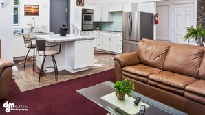 Een moderne keuken met witte kasten, roestvrijstalen apparatuur en een centraal keukenblok. Er staat een bruine leren bank en een glazen salontafel met een plant.