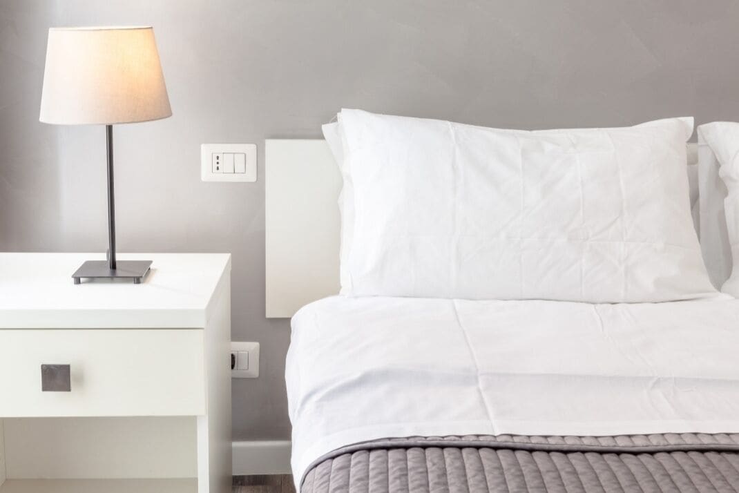 A neatly arranged hotel room with a white bed, a bedside table with a lamp, and a power outlet on the wall.