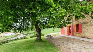Ein von Bäumen gesäumter Innenhof mit Picknicktisch und einem Steinhaus mit roten Fensterläden.