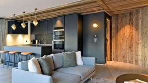 A modern living room with a grey sofa, a coffee table, and a kitchen area with dark cabinetry and wooden accents. - Luxury Apartment with Spa Access Near Slopes - The Allues (Méribel)