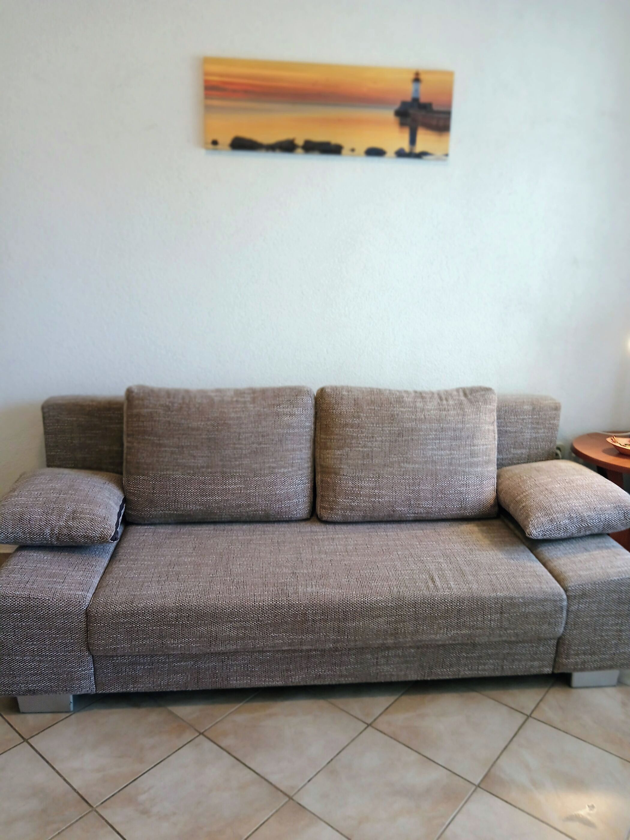 A grey sofa with a textured fabric, a small side table, and a painting of a lighthouse on the wall.