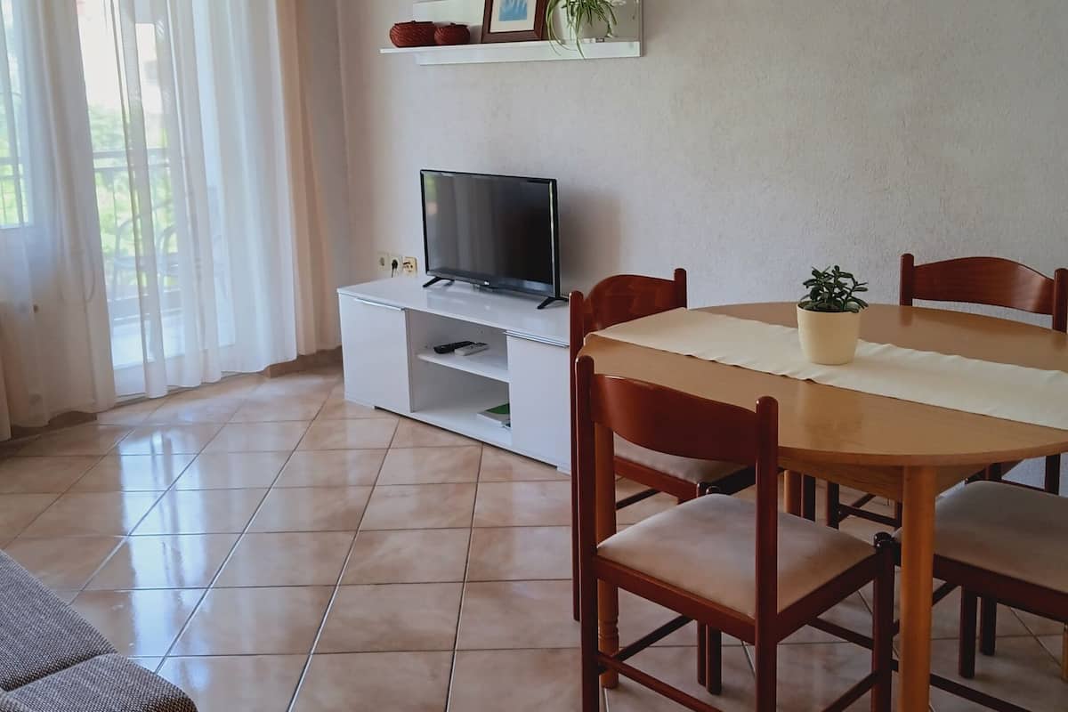 Un salon avec un téléviseur posé sur un meuble blanc, une table à manger avec des chaises, un canapé et une fenêtre avec des rideaux.