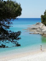 Una playa con agua turquesa cristalina, costa rocosa y una persona caminando sobre la arena.