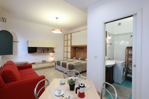 Een compacte hotelkamer met kitchenette, een zithoek met tafel en stoelen, een rode bank, een bed met houten hoofdeinde en een badkamer met wastafel en wasmachine.